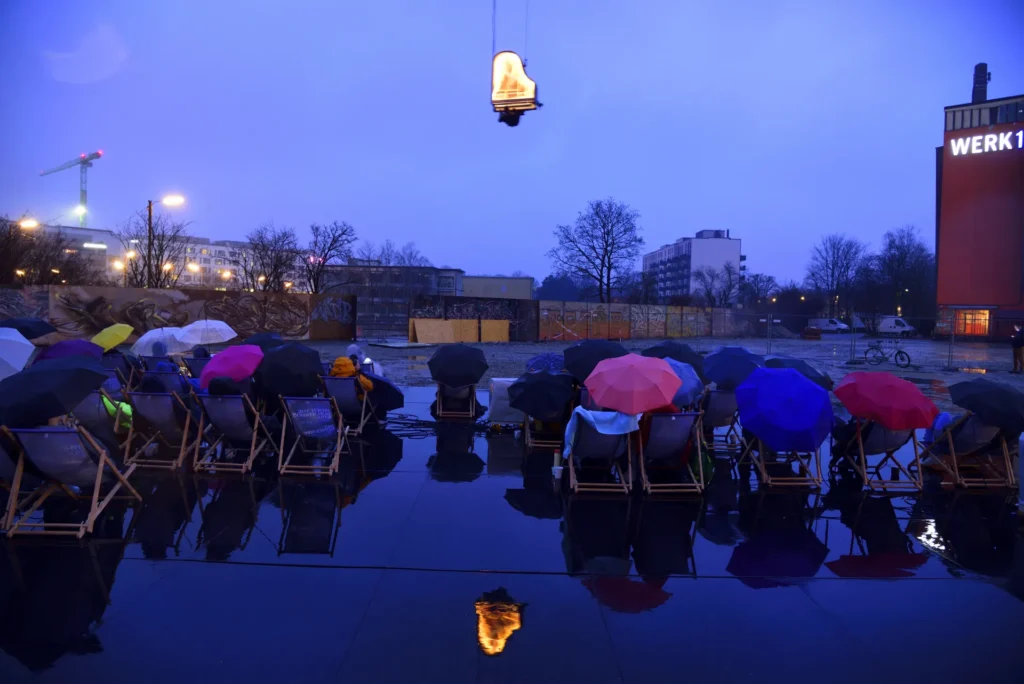 Alain Roche spielt einen Flügel am Kran hängend in München am 17.2.2024. Im Vordergrund Zuschauer in Liegenstühlen mit Regenschirmen.